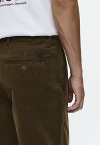 Bruine corduroybroek met een ribbelstructuur, voorzien van een achterzak met een knoop en geborduurd "W" logo detail.