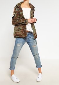 Kamouflagejacka med huva, prydd med militärfickor, över en vit topp. Ljusblå rivna jeans och vita sneakers fulländar looken.