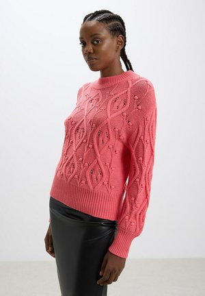 Maglione rosa lavorato a maglia con un design strutturato, caratterizzato da motivi a treccia e dettagli in pizzo, completato da polsini e bordo a costine.