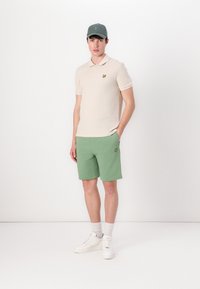Beige poloko s krátkým rukávem a malým emblémem v kombinaci se zelenými šortkami a bílými teniskami se světle šedými ponožkami.