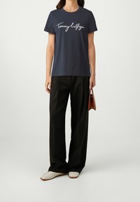 Marineblaues Baumwoll-T-Shirt mit weißem "Tommy Hilfiger"-Logo, kombiniert mit schwarzen weiten Hosen und cremefarbenen Slip-On-Schuhen. Braune Handtasche in der Hand.