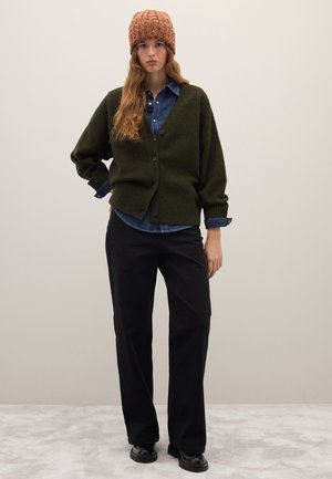 Cárdigan de punto verde oscuro con un corte holgado, pantalones negros y un gorro de punto naranja texturizado sobre una camisa de mezclilla azul.