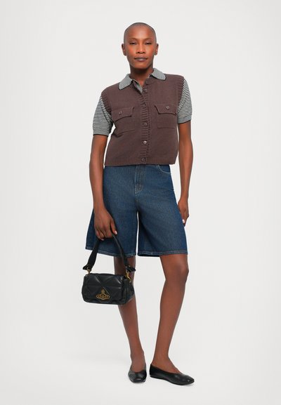 Top senza maniche in maglia marrone con colletto a righe, due tasche frontali, abbinato a pantaloni corti in denim blu e una piccola borsa nera trapuntata.