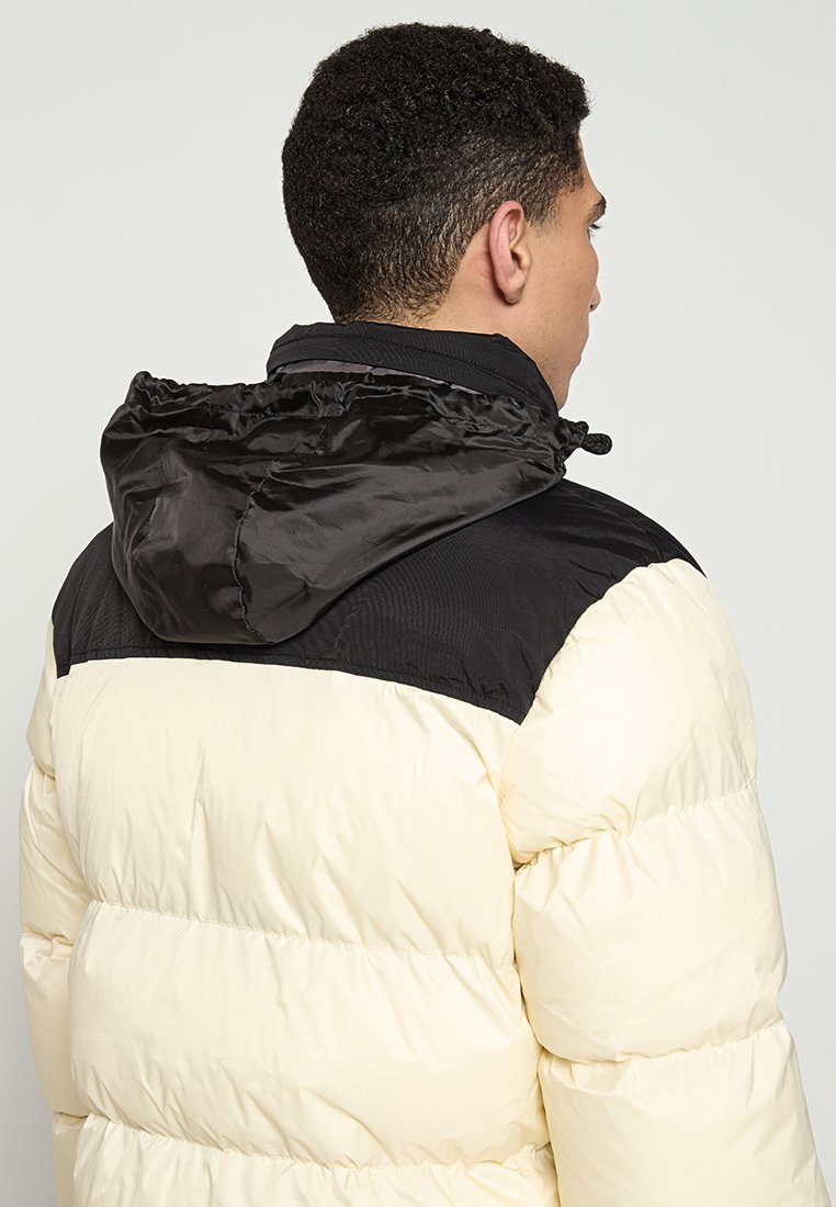 Mann mit kurzen lockigen Haaren, der eine cremefarbene und schwarze Steppjacke mit einer schwarzen Kapuze trägt, von hinten gezeigt und leicht zur Seite gedreht.