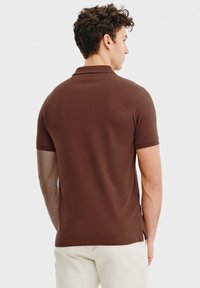 Brązowa koszulka polo z miękkiego materiału, z krótkimi rękawami, posiadająca standardowy kołnierzyk i gładkie linie. Widoczne przeszycia wzdłuż dołu.