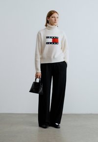 Pull en maille blanc avec col roulé et logo "TOMMY JEANS" multicolore, associé à un pantalon large noir et un petit sac à main noir.