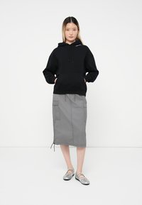 Vrouw die met haar handen in de zak staat, gekleed in een zwarte hoodie, een grijze cargorok en zilveren ballerina's, tegen een witte achtergrond.