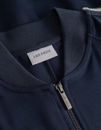 Μπλε ζακέτα με φερμουάρ και ριγωτό γιακά. Διαθέτει ασημένιο φερμουάρ και ετικέτα που λέει "LES DEUX." Το ύφασμα φαίνεται λείο και ανθεκτικό.