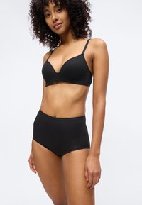 Γυναίκα με σγουρά μαλλιά, φοράει μαύρο άνετο σουτιέν χωρίς ραφές και μαύρο ψηλόμεσο εσώρουχο, στέκεται μπροστά σε λευκό φόντο.