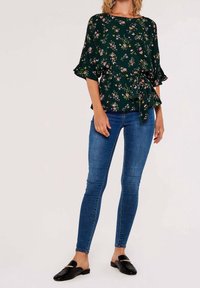 Blouse verte à motif floral avec manches à volants et taille nouée, assortie à un jean skinny bleu et des mocassins noirs.