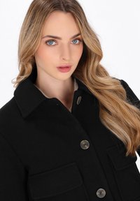 Ung kvinde med langt blond hår iført en sort knapcoats, vender ansigtet fremad med et neutralt udtryk og let makeup.