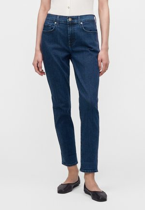 Slim fit mörkblå jeans, använda med svarta platta skor och en vit ärmlös topp, stående mot en enkel ljus bakgrund.