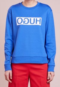 Niebieska bluza z miękkiego materiału, z białym prostokątnym logo "HUGO" na przodzie. Długie rękawy i luźny krój.