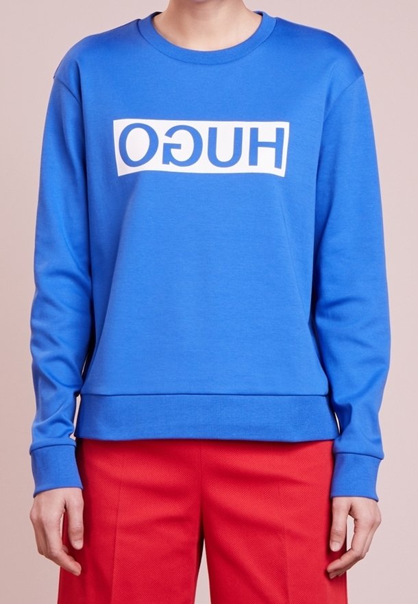 Niebieska bluza z miękkiego materiału, z białym prostokątnym logo "HUGO" na przodzie. Długie rękawy i luźny krój.