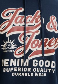 Sweat à capuche bleu marine avec texte rouge et blanc « Jack & Jones » et phrases « Denim Good », « Superior Quality » et « Durable Wear », plus un logo soleil avec « Since 1990 .»