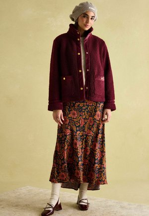 Burgunderfarbener strukturierter Blazer mit goldenen Knöpfen, kombiniert mit einem dunkel gemusterten Rock, cremefarbene Socken und braunen Mary Jane Schuhen.
