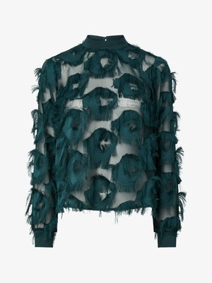 Blouse en tulle teal avec des motifs circulaires texturés et effilochés, col haut et manches longues avec un ourlet droit. Fermeture au niveau de la nuque par un trou clé.