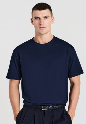 Giovane uomo con capelli corti castani, che indossa una semplice maglietta blu navy infilata nei pantaloni neri con una cintura nera, in piedi con le mani nelle tasche.