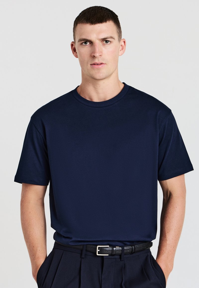 Giovane uomo con capelli corti castani, che indossa una semplice maglietta blu navy infilata nei pantaloni neri con una cintura nera, in piedi con le mani nelle tasche.