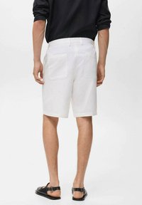 Pantalones cortos de algodón blancos con corte recto y dos bolsillos traseros, combinados con sandalias negras, vistos desde atrás.
