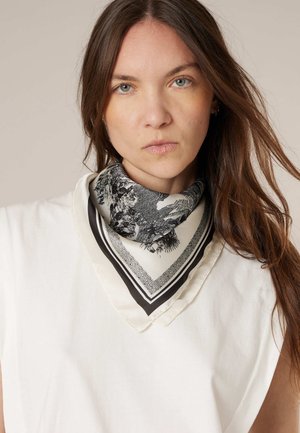 Femme aux longs cheveux bruns portant un haut blanc et une écharpe à motif noir et blanc nouée autour du cou, fond neutre.