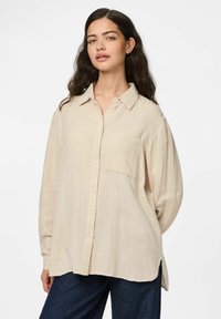 Camicia beige con bottoni, realizzata in tessuto strutturato, dotata di una tasca sul petto e maniche lunghe con polsini. L'orlo è leggermente più lungo sul retro.