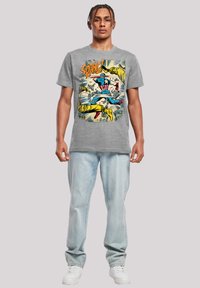 Grå bomull t-skjorte med et fargerikt grafisk trykk av en superhelt. Modellen har på seg lyseblå jeans og hvite sneakers.