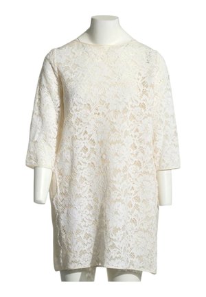 Blouse blanche en dentelle florale à manches longues avec un col rond, présentée sur un mannequin sans tête sur fond blanc.