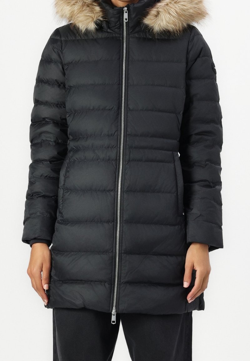 Manteau matelassé noir avec fermeture éclair à l'avant et capuche bordée de fourrure porté par une personne visible du cou jusqu'à mi-cuisse.