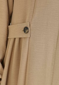 Beige stof med en tekstureret overflade, der har et plisseret design og en stor, rund knapdetalje på venstre side.