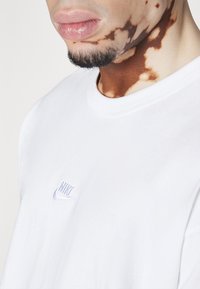 White cotton t-shirt featuring a small blue Nike logo on the chest. The fabric has a smooth texture, and the neckline is a classic crew style.