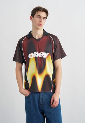 Ung mand med kort hår iført en sort og orange skjorte med flammemønster og teksten "obey", parret med løse blå jeans, stående mod en hvid baggrund.