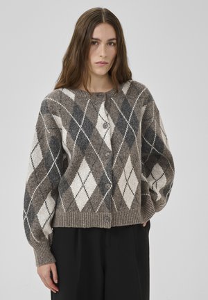 Mujer con cabello largo y castaño que lleva un cárdigan de punto con botones, de patrón romboidal gris y blanco, y pantalones negros, sobre un fondo liso.