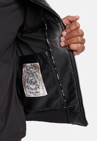 Interior de chaqueta de cuero negro con una etiqueta de color crema que presenta un logotipo y "Est. 1981". Detalle de cremallera visible a lo largo del borde, textura suave.