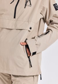 Beige pull-over jack gemaakt van lichtgewicht stof, met een grote voorkant zak, een ritssluiting zijzak en logodetails op de borst en mouw.