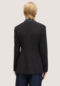 Blazer negro entallado que presenta una silueta ajustada, solapas pronunciadas y sutiles pliegues en la espalda. La tela parece suave con una forma estructurada.