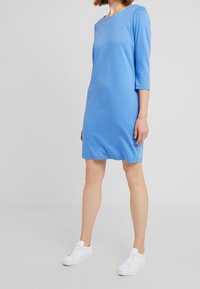 Blauwe jurk tot op de knie met 3/4 mouwen. Gladde textuur en ronde halslijn, gecombineerd met witte sneakers voor een casual uitstraling.