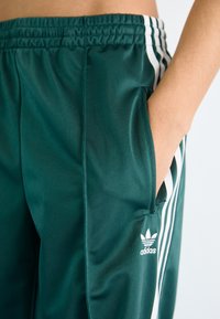 Green polyester athletic pants feature a drawstring waistband, side pockets, and white three-stripe accents along the sides. Adidas logo at hip.