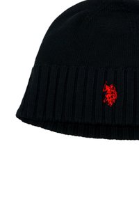 Căciulă tricotată neagră cu manșetă ribbed. Prezintă un logo roșu proeminent al unui jucător de polo brodat pe față. Textură moale.