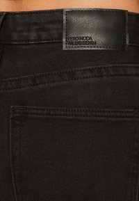 Jeans de mezclilla negros con un parche de cuero que dice "VERO MODA WE DO DENIM" en relieve, que muestran una textura suave y detalles de costura sencilla.