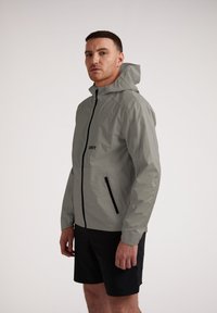 Giacca impermeabile grigia chiara con cappuccio, cerniera nera e tasche, caratterizzata da un design semplice e una superficie liscia, indossata sopra pantaloni corti neri.