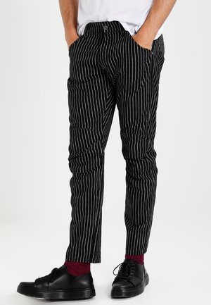 Mann trägt schwarze Hose mit weißen vertikalen gepunkteten Streifen, burgunderrote Socken, schwarze Lederschuhe und ein weißes Hemd, die Hände in den Taschen.