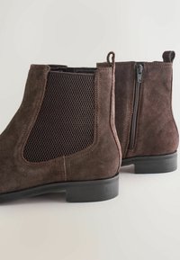 Botas de tobillo de ante marrón con paneles de malla, cierre con cremallera lateral y un bajo tacón de goma negra. Presentan una textura suave y un detallado acabado en costuras.