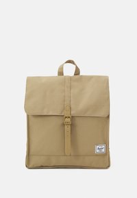 Khaki canvas backpack with a flap closure, front buckle strap, top handle, and a logo patch. Smooth texture and rectangular shape.