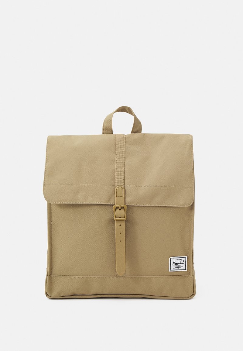 Khaki canvas backpack with a flap closure, front buckle strap, top handle, and a logo patch. Smooth texture and rectangular shape.