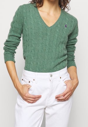 Femme portant un pull vert tricoté en torsades à col en V et un pantalon blanc taille haute, les mains dans les poches, sur un fond uni.