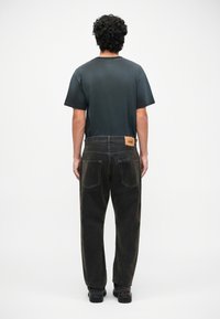 Jeanși negri din denim cu un finisaj decolorat, având o eticheta pătrată pe spate și două buzunare pe spate. Asortați cu o cămașă verde închis.