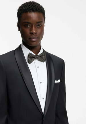 Man wearing black tuxedo with satin lapels, white pleated shirt with studs, patterned black bow tie, and white pocket square against white background.
