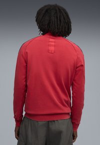 Rød sweatshirt med lange ærmer, ribbede manchetter og rund halsudskæring; har en lille flap bagpå og sorte detaljer på skuldrene.
