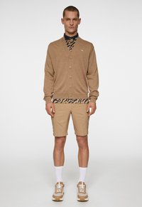 Brun cardigan med knappar, lagd över en marinblå skjorta med leopardskrift, ihop med beige shorts och sneakers. Vita strumpor synliga.